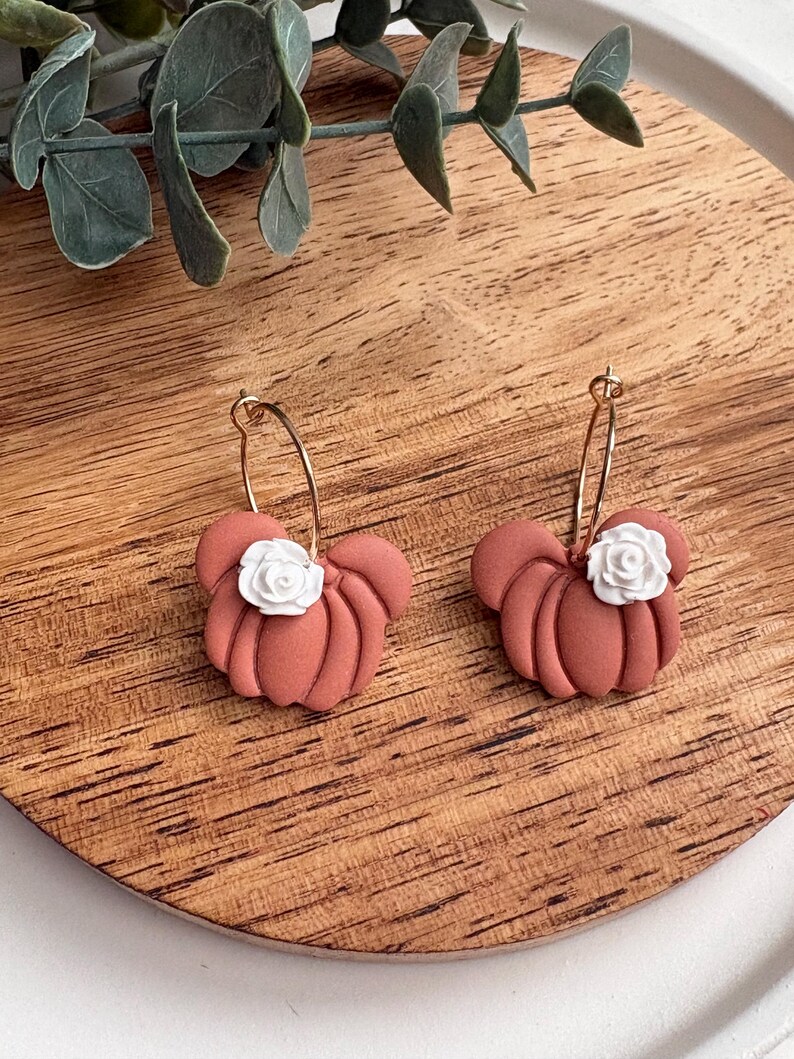 Puede incluir: Un par de pendientes con forma de calabaza de color terracota con detalles de rosas blancas. Los pendientes est&aacute;n sujetos a cierres de aro dorados. Los pendientes se exhiben sobre una superficie de madera.