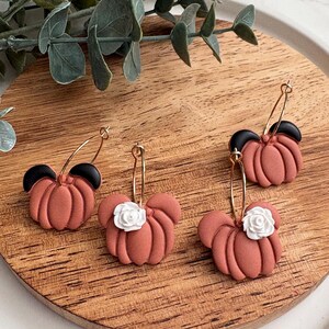 Puede incluir: Cuatro pares de pendientes con forma de calabaza de color terracota con aros dorados. Dos pares tienen detalles negros y dos tienen detalles de flores blancas. Los pendientes se exhiben sobre una superficie de madera.