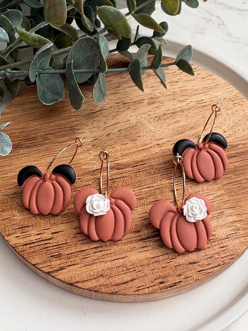 Puede incluir: Cuatro pares de pendientes con forma de calabaza de color terracota con aros dorados. Dos pares tienen detalles negros y dos tienen detalles de flores blancas. Los pendientes se exhiben sobre una superficie de madera.