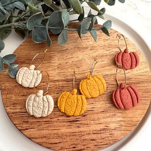 May include: A collection of pumpkin-shaped earrings in cream, yellow, and terracotta colors. The earrings are on gold hoops and feature detailed embossed designs. The earrings are displayed on a wooden surface, with greenery in the background.