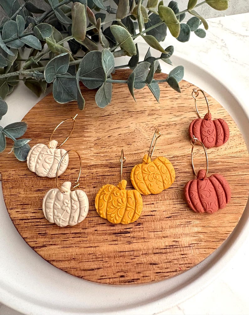 May include: A collection of pumpkin-shaped earrings in cream, yellow, and terracotta colors. The earrings are on gold hoops and feature detailed embossed designs. The earrings are displayed on a wooden surface, with greenery in the background.