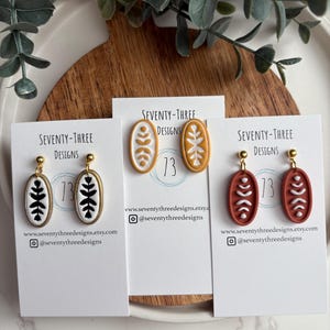 May include: Three pairs of oval-shaped earrings with gold-colored posts. Each earring features a different design: black and white, white and yellow, and white and rust-colored. The earrings are displayed on white cards with the text "SEVENTY-THREE DESIGNS".