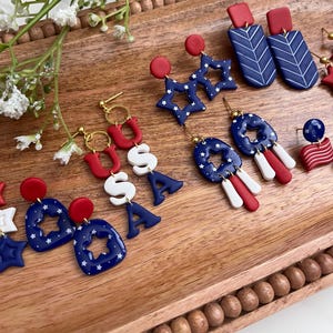 May include: A selection of red, white, and blue earrings with patriotic themes. The designs include star shapes, the letters "USA", and geometric patterns. The earrings are presented on a wooden tray, highlighting their festive colours and details.
