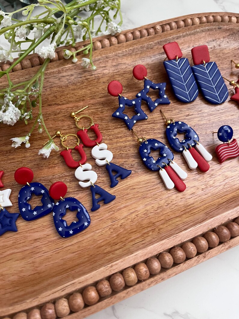 May include: A selection of red, white, and blue earrings with patriotic themes. The designs include star shapes, the letters "USA", and geometric patterns. The earrings are presented on a wooden tray, highlighting their festive colours and details.