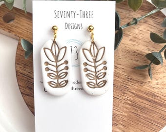 May include: A pair of white oval earrings with a gold floral design. The earrings are hanging from gold-colored posts. The earrings are displayed on a white card with the text "SEVENTY-THREE DESIGNS".