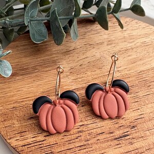 Puede incluir: Pendientes hechos a mano con un dise&ntilde;o de calabaza. Los pendientes presentan calabazas de color terracota con la parte superior negra, suspendidas de aros dorados. Los pendientes se exhiben sobre una superficie de madera.