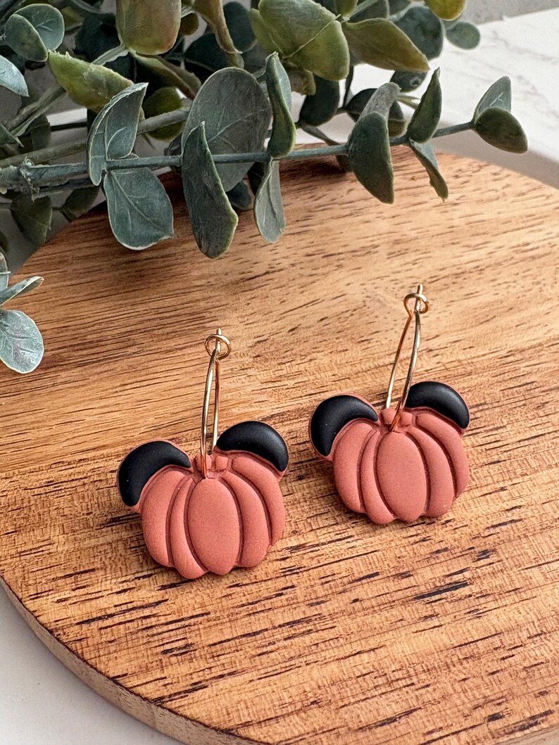 Puede incluir: Pendientes hechos a mano con un dise&ntilde;o de calabaza. Los pendientes presentan calabazas de color terracota con la parte superior negra, suspendidas de aros dorados. Los pendientes se exhiben sobre una superficie de madera.