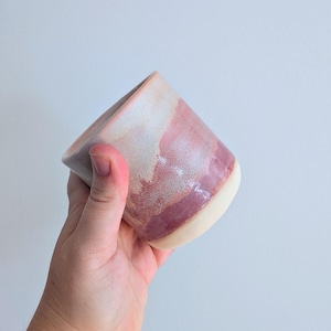 May include: A ceramic tumbler featuring a smooth, cylindrical shape with a tapered base. The exterior displays a blended glaze transition from a creamy off-white at the bottom to a deep, dusty rose pink at the top, with a glossy finish.