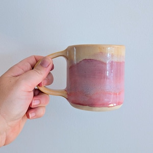 Peut inclure: Une tasse en céramique avec une finition brillante, présentant une partie supérieure de couleur crème et une partie inférieure aux tons mélangés de rose et de mauve. La tasse possède une anse incurvée et un corps arrondi à l'aspect artisanal.