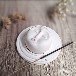 May include: A white ceramic incense holder featuring a smiling moon face design. The holder sits on a circular base with concentric rings. An incense stick rests on the base. The holder is a decorative item.