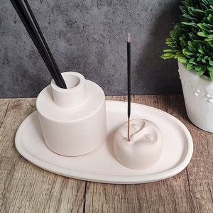 May include: A light pink ceramic incense holder set. The set includes a cylindrical diffuser with black reeds, a round incense holder, and an oval tray. A lit incense stick is in the round holder. A small potted plant is in the background.