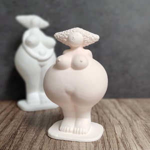 May include: A light pink ceramic sculpture of a stylized female form with a rounded belly and small feet. The figure has a small head with curly hair and is standing on a circular base. Another white sculpture is in the background.