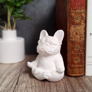 May include: A light pink French bulldog figurine in a seated yoga pose. The sculpture is on a wooden surface, with a white vase and books in the background. The figurine has detailed facial features and is a decorative item.