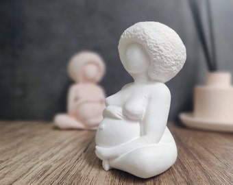 Op de afbeelding: Een witte, minimalistische sculptuur van een zwangere persoon in zittende positie. De figuur heeft een groot, getextureerd afro-kapsel en houdt haar buik vast. Een andere, kleinere, roze sculptuur is wazig op de achtergrond. De sculpturen staan op een houten oppervlak.