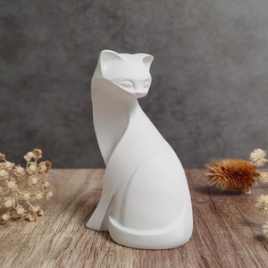 May include: A white cat figurine with a sleek, minimalist design. The sculpture is sitting on a wooden surface, with dried flowers and a dark gray background. The cat is in a seated position, with its head tilted slightly downward.