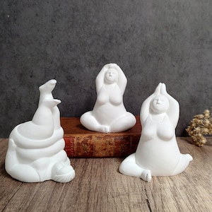 May include: Three white yoga figurines in different poses are displayed on a wooden surface. One is upside down, another is seated with arms raised, and the third is in a seated prayer position. The background is a dark gray wall.