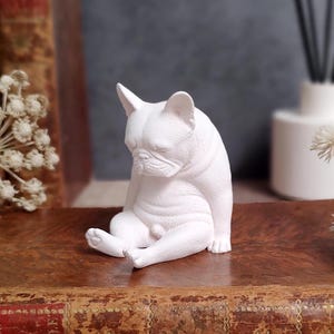 May include: A white ceramic figurine of a French Bulldog sitting with its head down. The dog is sitting on a brown surface. The figurine has a smooth texture and is a solid white color.