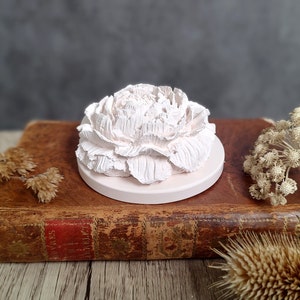 May include: A white ceramic flower diffuser sits on top of an old, brown book. The flower is detailed and realistic, with many petals.