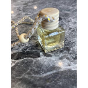 Puede incluir: Una pequeña botella cuadrada de vidrio llena de un líquido amarillo, coronada con una tapa de madera clara. Se adjunta una cuerda decorativa con detalles dorados y una cuenta de madera. La botella está diseñada para aromaterapia o fragancia.