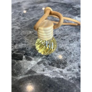 Puede incluir: Una pequeña botella de vidrio transparente con un diseño acanalado, llena de líquido amarillo. La botella tiene una tapa de madera de color claro y una cuerda marrón para colgar. Probablemente sea un ambientador o un difusor de aceites esenciales.
