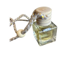 Puede incluir: Una pequeña botella cuadrada de vidrio transparente con una tapa de madera de color claro. La botella contiene un líquido amarillo y está unida a una cuerda beige y dorada con una cuenta de madera. Probablemente sea un ambientador o difusor de fragancia.