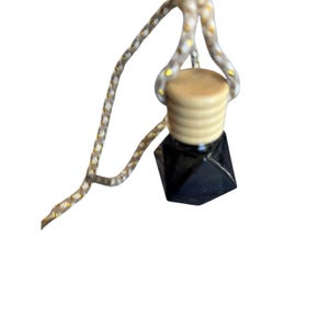 Puede incluir: Una pequeña botella negra de forma geométrica con una tapa beige está suspendida por un cordón blanco y dorado. La tapa de la botella es cilíndrica con estrías horizontales. El cordón está en bucle en la parte superior.