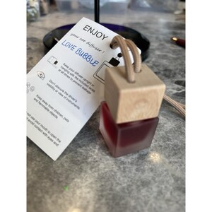 Puede incluir: Un ambientador para coche con una parte superior de madera y una base roja de forma cuadrada. El producto está etiquetado como "LOVE BUBBLE" e incluye una etiqueta con instrucciones. Se adjunta una cuerda beige para colgar.