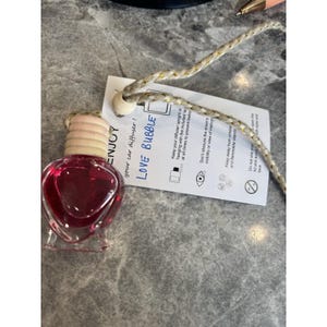 Op de afbeelding: Een hartvormige auto-geurverspreider gevuld met dieprode vloeistof. De fles heeft een houten dop en een label met "LOVE BUBBLE" en "ENJOY your car diffuser!". Hij hangt aan een goud-wit koord.
