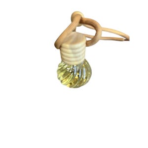 Puede incluir: Una pequeña botella de vidrio transparente con un diseño acanalado, llena de un líquido amarillo. La botella tiene una tapa de madera y está unida a un cordón marrón claro, probablemente para colgar. Parece ser un ambientador o difusor de fragancia.