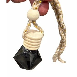 May include: A black, faceted glass bottle with a wooden cap and a beige rope with gold accents. The bottle is suspended by the rope, which is held by a person's fingers.
