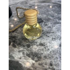 Puede incluir: Una pequeña botella de vidrio transparente llena de un líquido amarillo, coronada con una tapa de madera. La botella está suspendida por una cuerda dorada. El producto parece ser una fragancia o aceite esencial, para aromaterapia o decoración.