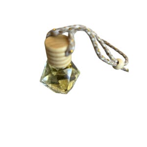 Puede incluir: Pequeña botella de vidrio transparente en forma de diamante, llena de líquido amarillo. Tiene una tapa de madera clara y un cordón beige con detalles dorados para colgar. La botella está diseñada para contener fragancia.