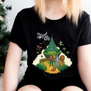 May include: Black t-shirt with a vibrant illustration from The Wizard of Oz. The design showcases the Emerald City, the yellow brick road, and characters such as the Tin Man and the Cowardly Lion. The text "The Wizard of Oz" is included.