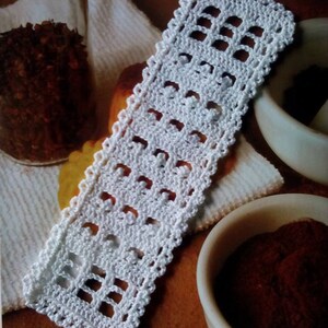 Op de afbeelding: Witte gehaakte bladwijzer met een vierkant patroon. De bladwijzer ligt op een bruin oppervlak met een witte handdoek en een kleine kom met bruin poeder.