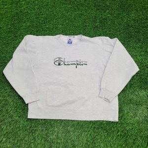 May include: A light gray Champion sweatshirt with a crew neck. The Champion logo is embroidered in green and gray on the front. The sweatshirt is made of a soft, comfortable material.