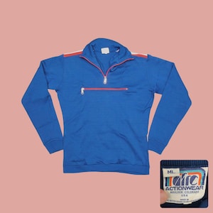May include: A blue vintage Actionwear pullover with a red and white stripe detail on the shoulders and a red zipper. The pullover has a small chest pocket with a red zipper and a tag that reads "Actionwear Boulder, Colorado USA".