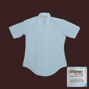 May include: A light blue short-sleeved dress shirt with a pointed collar and button-down front. The shirt has a curved hem and a label that reads "creations by Jason Clarke". The fabric is 65% polyester and 35% cotton.