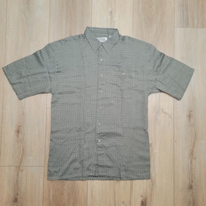 May include: A short-sleeved button-down shirt in a muted green hue, featuring a subtle, repeating geometric pattern. The shirt has a classic collar, a chest pocket, and is buttoned up. The shirt is laid flat on a wooden surface.