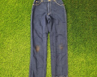 May include: Dark blue denim jeans with a classic five-pocket design. The jeans have a high waist and a straight leg cut. There are two brown stains on the front of the jeans. The jeans are laid out on a green surface.