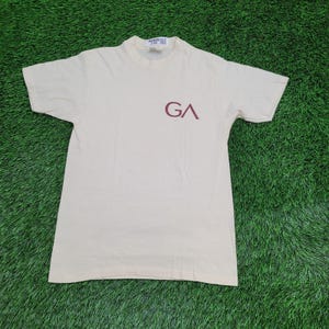 May include: Cream-colored t-shirt with short sleeves and a crew neck. The shirt features the maroon letters "GA" on the front. The t-shirt is laid flat on a green surface.