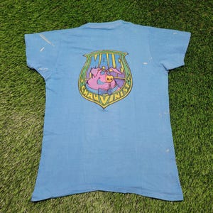 May include: Light blue t-shirt with a graphic on the back featuring a pig wearing a bow tie and holding a cigarette. The graphic is in a shield shape with the words "Male Chauvinist" around the pig.