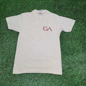 May include: A cream-colored short-sleeve t-shirt featuring a maroon graphic on the chest with the letters GA. The shirt has a crew neckline and a small white tag at the inner collar with the number 86409 printed in black text.