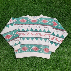 May include: White crew neck sweatshirt with a Southwestern geometric pattern in teal, red, and black. The long-sleeved top features a ribbed collar, cuffs, and hem. The design includes zigzags, triangles, and stylized shapes.