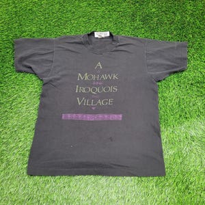 May include: A dark gray t-shirt with the text "A Mohawk Iroquois Village" in green and purple. The shirt is laid out on a green grassy surface.