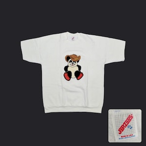 May include: A white short-sleeve t-shirt featuring a graphic of a panda wearing a brown cowboy hat and red shoes. The shirt includes a white tag with a red Jerzees logo, the size XL 46, and the text Made in U.S.A. printed on the fabric.