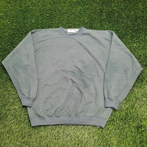 May include: A gray-green crew neck sweatshirt with long sleeves. The cuffs and hem are a darker shade of gray-green. The sweatshirt is lying on a green surface.