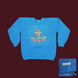 May include: A turquoise blue sweatshirt with a crew neck and long sleeves. The front features a colorful, abstract design with a globe-like shape and a plaid pattern. The sweatshirt is made of a soft material.