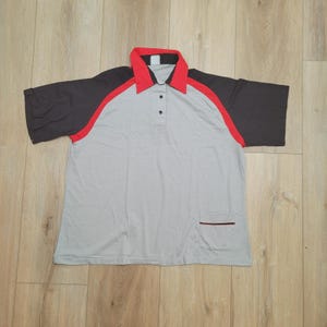 May include: A gray polo shirt with black sleeves and red accents. The shirt has a red collar and a small pocket on the lower left side. The shirt has three black buttons.