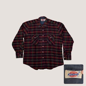 May include: A long-sleeved, button-down flannel shirt in a red, black, and green plaid pattern. The shirt has two chest pockets and a blue collar lining. The Dickies brand label is visible in the lower right corner.