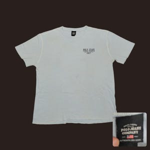 May include: A light gray short-sleeved t-shirt with a crew neckline. The chest features the text POLO JEANS RALPH LAUREN 1967 in dark lettering. A black interior neck label displays the text POLO JEANS COMPANY EST 1967 with an American flag icon.